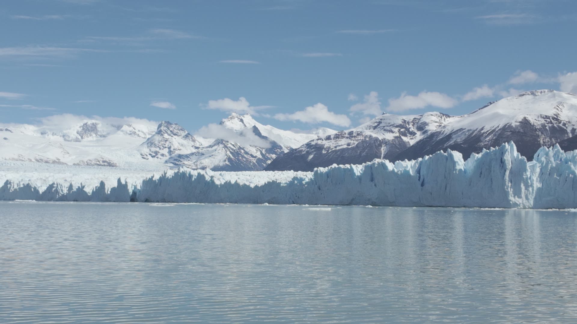 A glacier meets the water beneath snow-covered mountains and a blue sky.